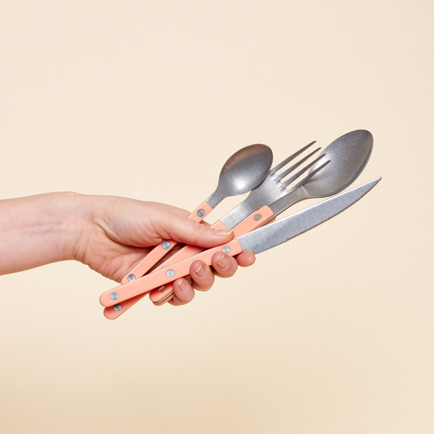 A hand holding a set of cutlery with peach-colored handles, including a knife, fork, and two spoons, against a neutral beige background.