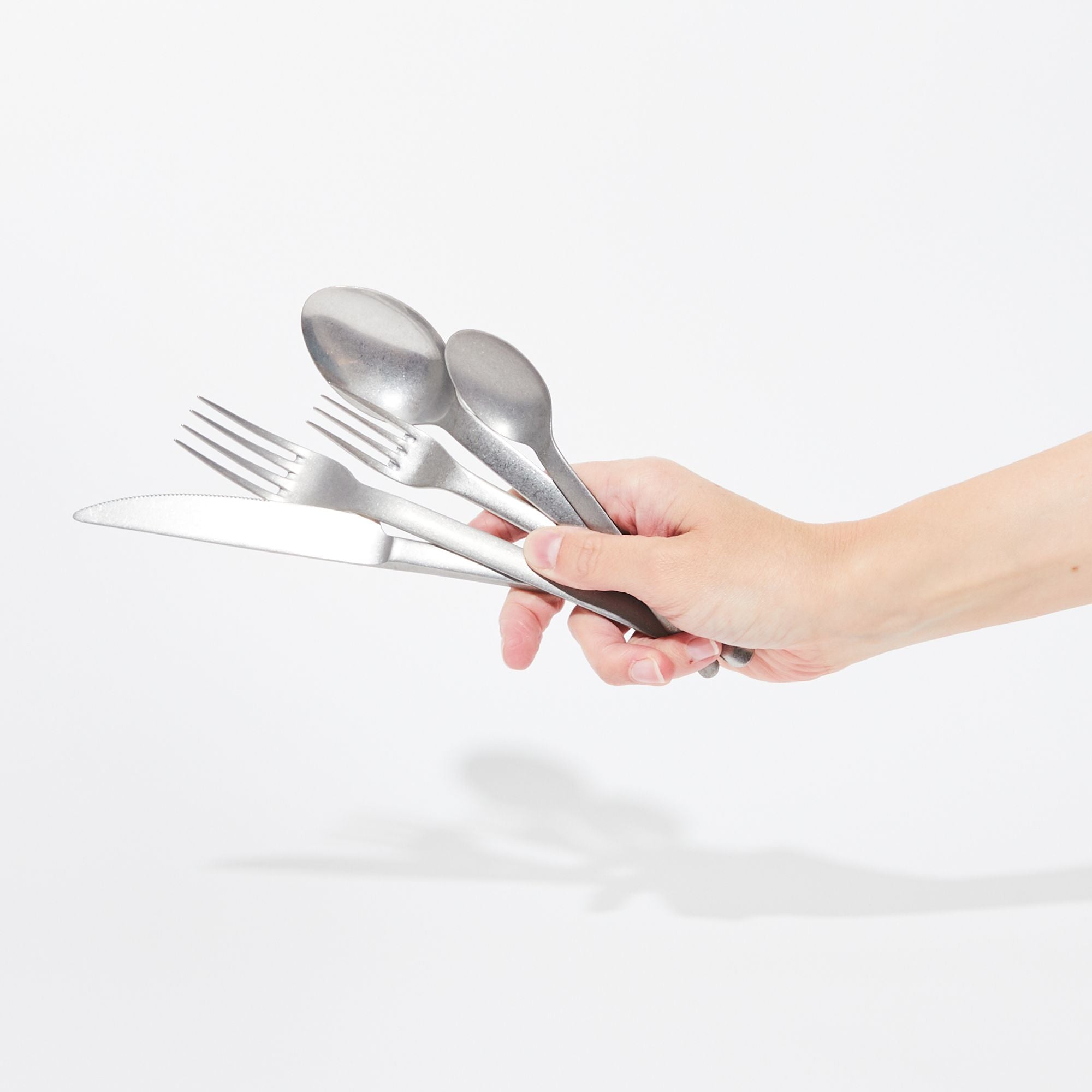 Hand holding stainless steel flatware (teaspoon, soup spoon, salad fork, dinner fork, and knife) in a vintage matte finish