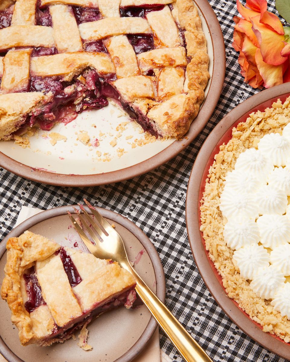 A lattice-top cherry pie with a slice removed sits on a black and white checkered cloth, next to a slice served on a plate with a gold fork. Adjacent is another dessert with a crumbly topping and whipped cream, alongside orange roses.