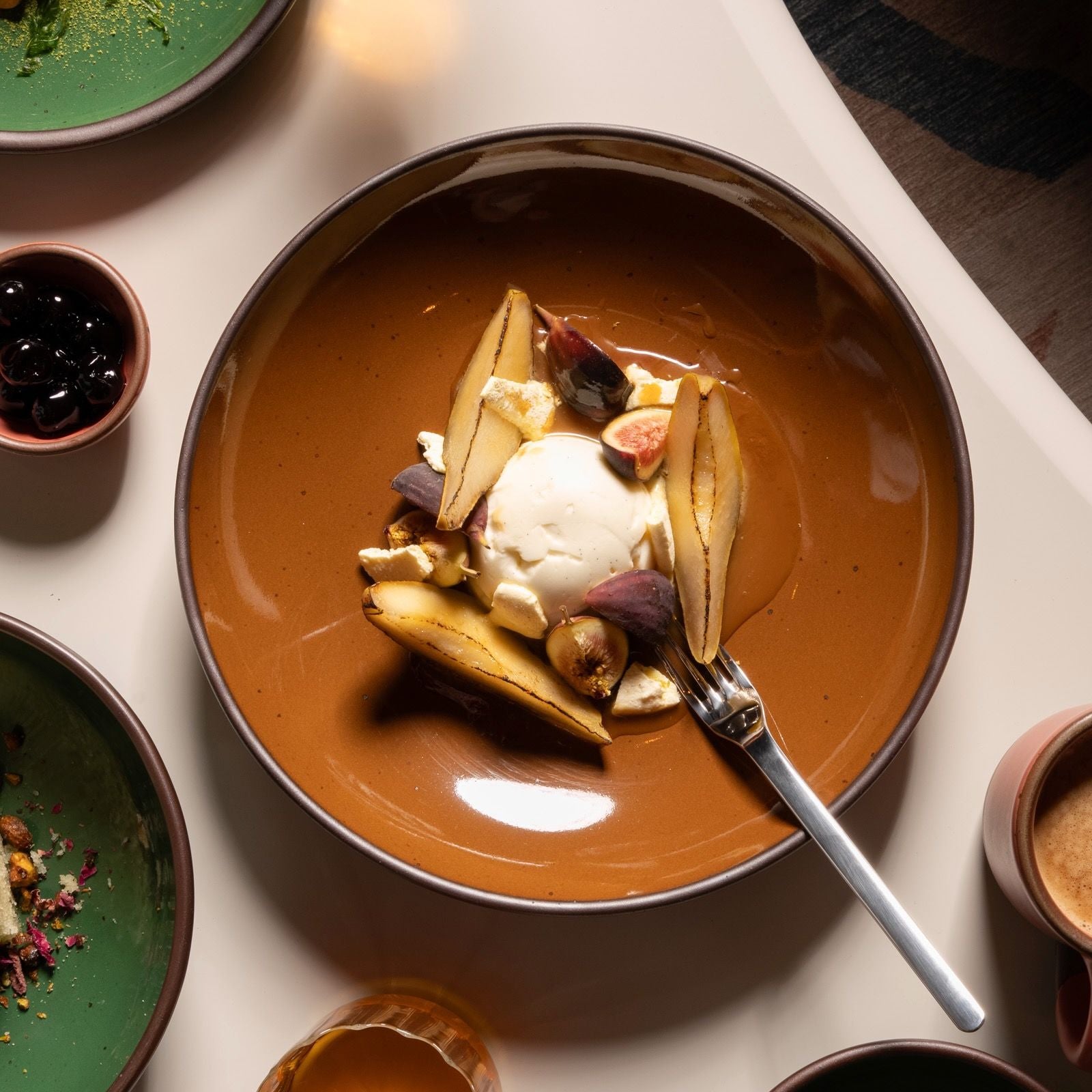A brown ceramic plate holds a dessert of poached pears, figs, and a scoop of vanilla ice cream, surrounded by green dishes and a cup of coffee.