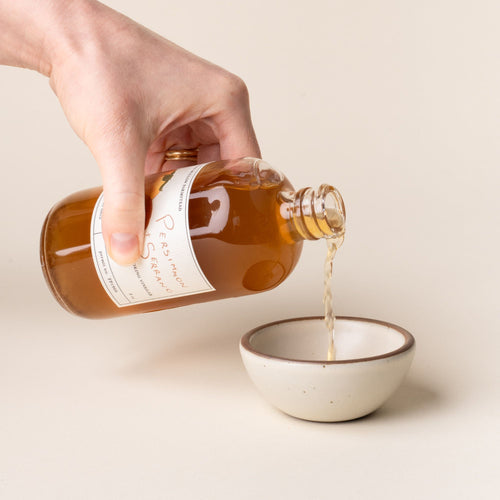 Hand holding a bottle of Persimmon + Serrano cooking vinegar, featuring hand-drawn persimmon illustrations on the label, pouring it into a small ceramic off-white bowl.
