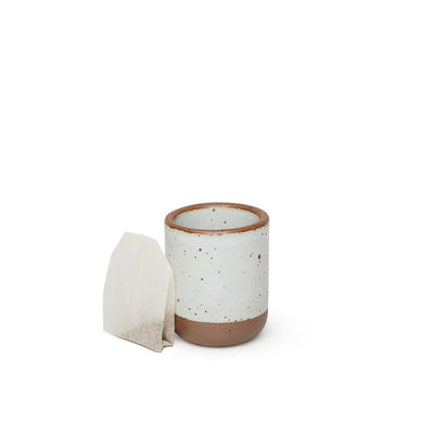 A teabag leaning next to a small, short ceramic mug cup in a cool white color featuring iron speckles and unglazed rim and bottom base.