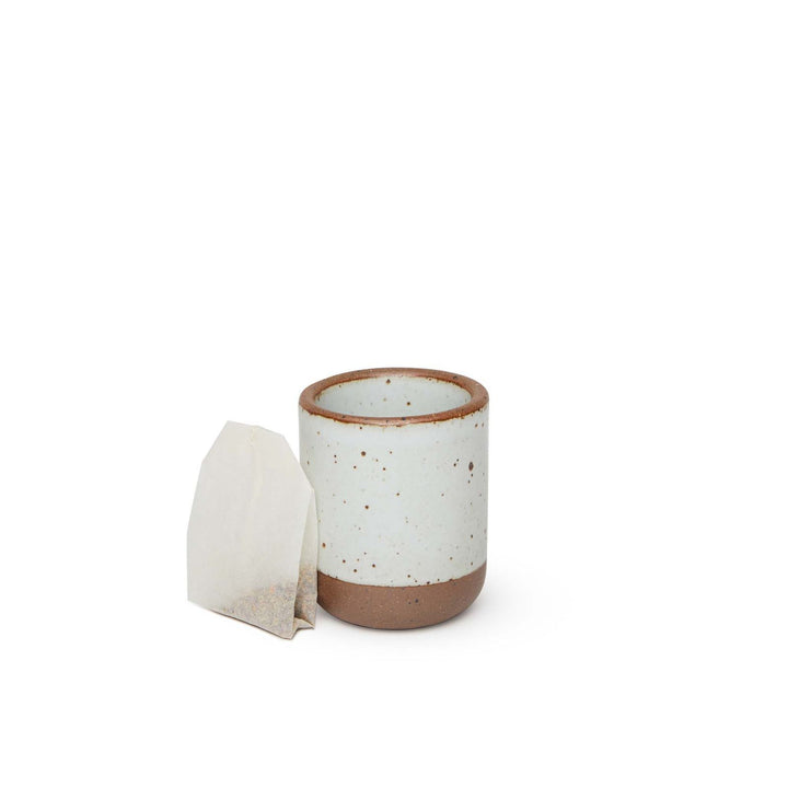 A teabag leaning next to a small, short ceramic mug cup in a cool white color featuring iron speckles and unglazed rim and bottom base.