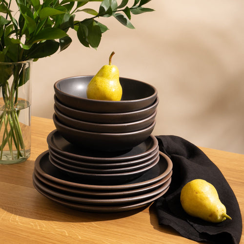4 ceramic dinner plates, 4 side plates, and 4 shallow dinner bowls in a graphite black color with iron speckles and unglazed rims are stacked all together on a table with pears.