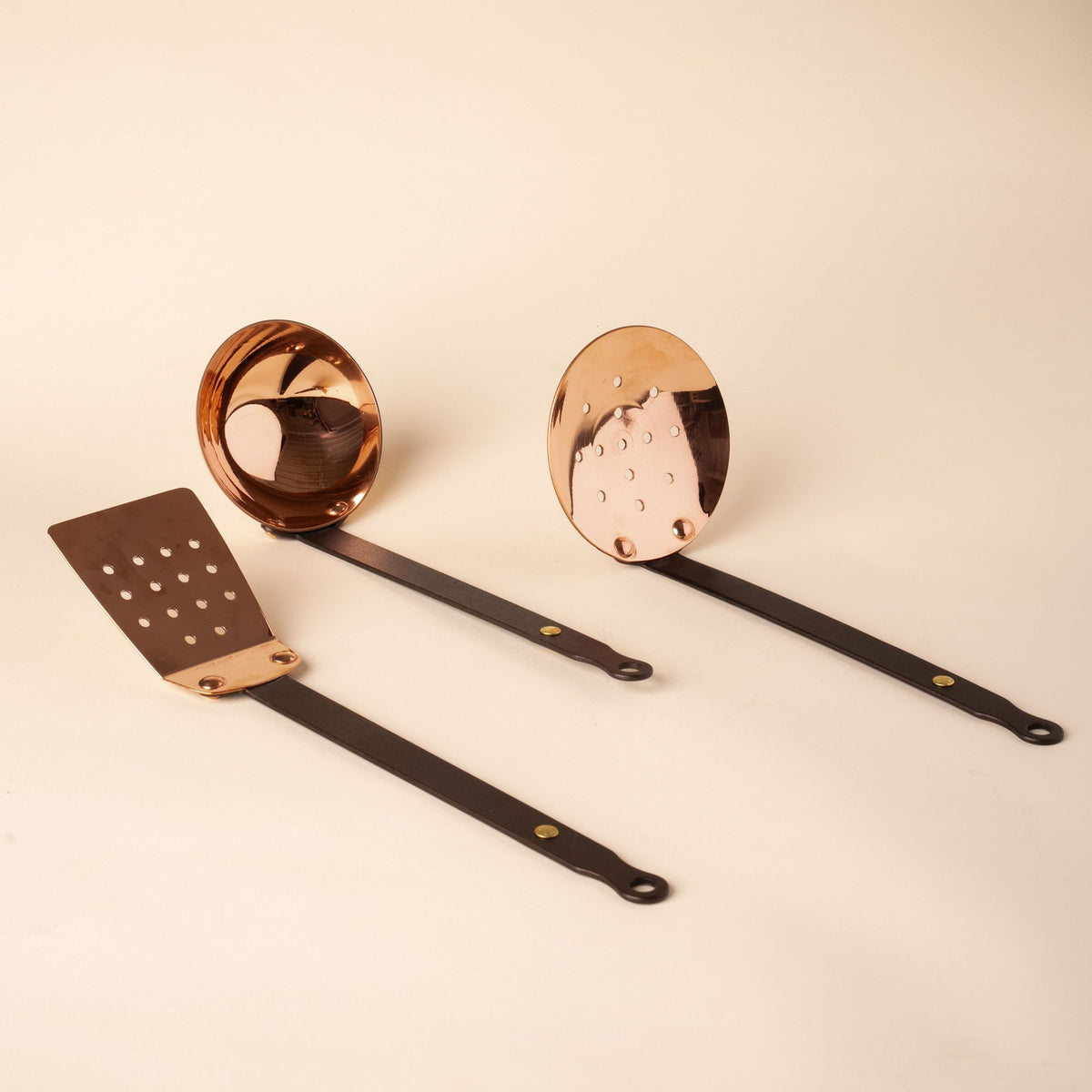Set of three copper kitchen utensils with black handles, including a spatula, ladle, and skimmer, displayed on a neutral background.