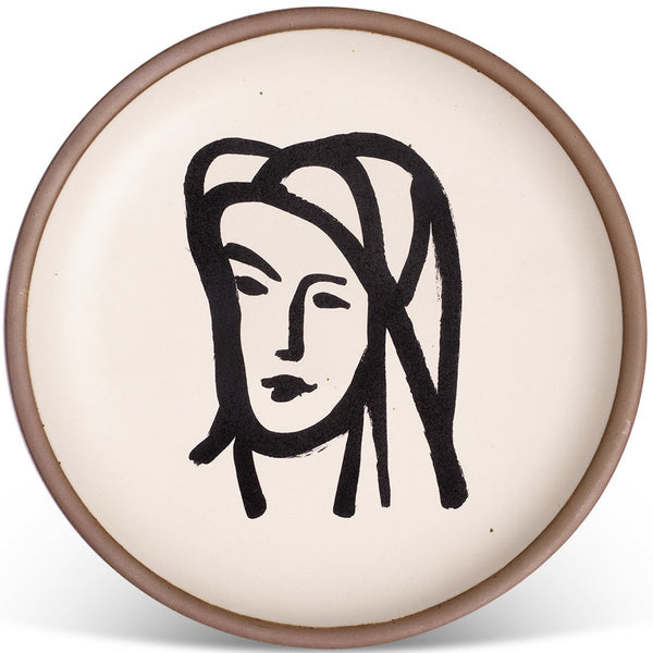 Round ceramic plate with a minimalist black line drawing of a woman's face on a cream background, bordered by a light brown rim.