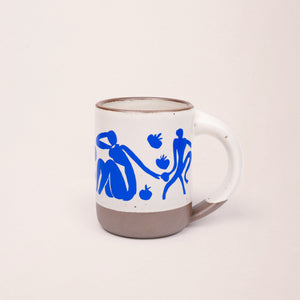 East Fork’s The Mug in a warm off white color with handle with a blue decal of Henri Matisse’s Femmes et singes, a bright blue, abstract cut-out figures of women sitting, monkeys, and pomegranate across the body of the mug