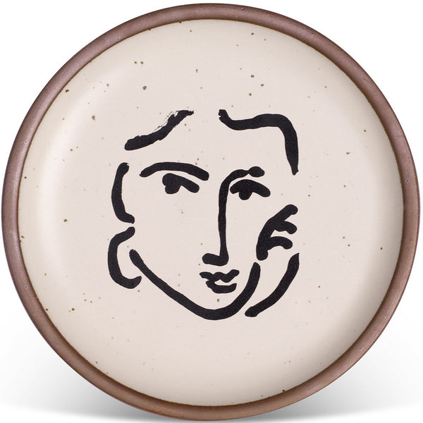 Round ceramic plate with a minimalist line drawing of a face in black on a speckled cream background, framed by a brown rim.