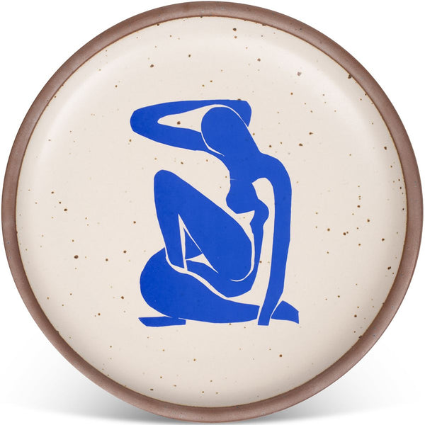 A round ceramic plate with a speckled cream surface, featuring a bold, abstract blue silhouette of a seated figure in the center, with a brown rim.