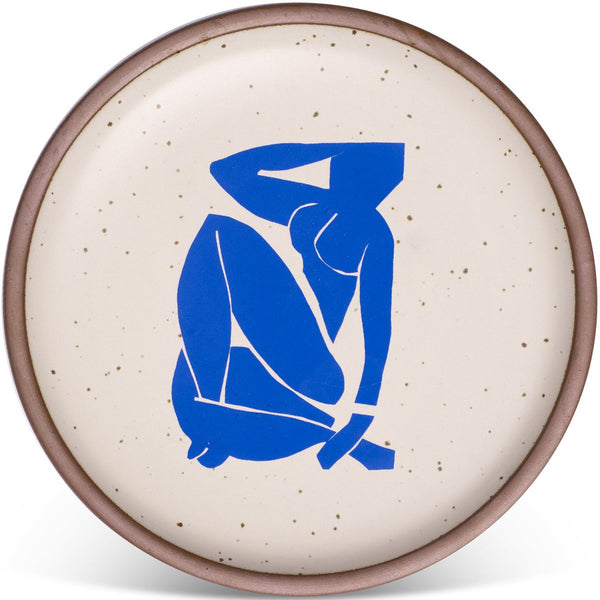 A round ceramic plate with a speckled beige surface, featuring a bold blue abstract figure design in the center. The plate has a subtle brown rim.