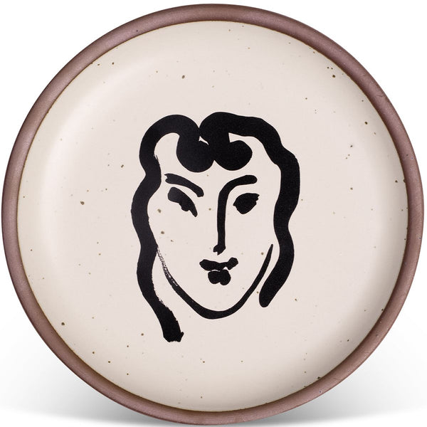 Ceramic plate with a speckled cream-colored surface featuring a minimalist black line drawing of a face in the center, bordered by a subtle earthy-toned rim.