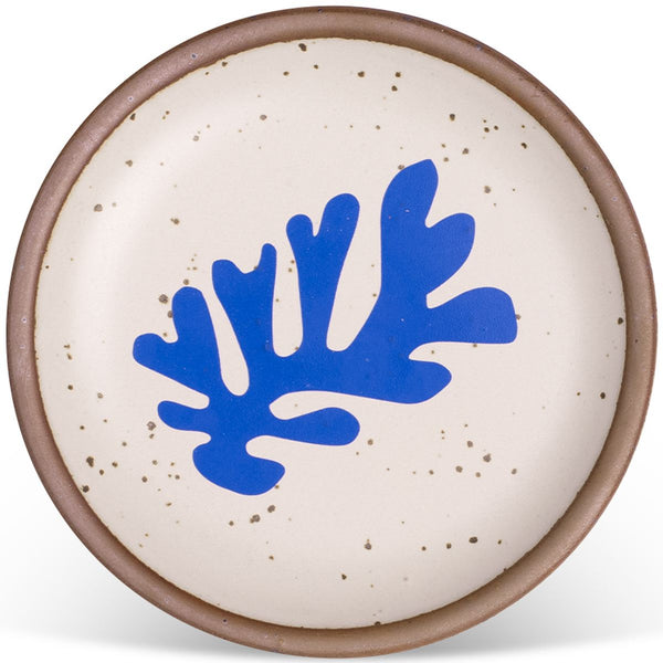 Round ceramic plate with a speckled cream surface, featuring a bold blue abstract leaf design in the center and a brown rim.