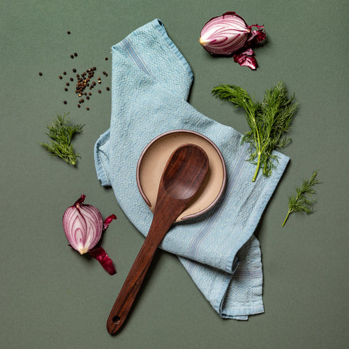 An overhead view of a ceramic spoon rest in a pale warm brown color with an unglazed rim, a blue kitchen towel, and a wooden kitchen spoon, surrounded by shallots, thyme, and peppercorns.