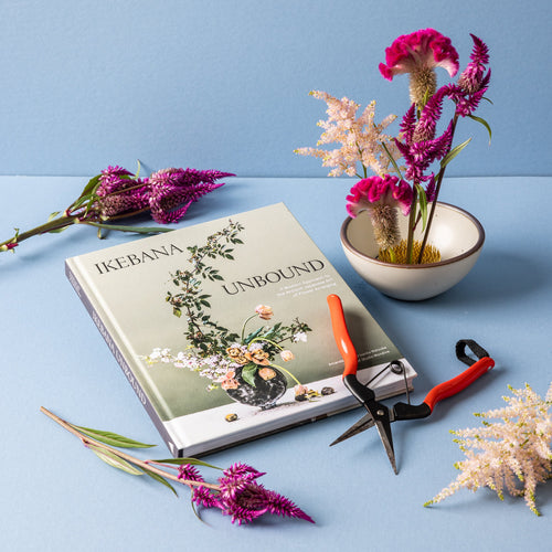 A book titled 'IKEBANA UNBOUND' lies open on a light blue surface, surrounded by pink and white flowers, red garden snips, and a small white bowl with flowers in a flower frog. 