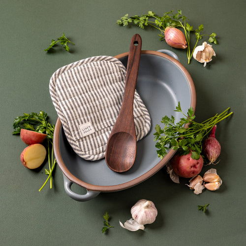 A styled overhead photo of a large oval baker in a blue-grey color with a wood spoon and striped pot holder with ingredients and herbs surrounding.