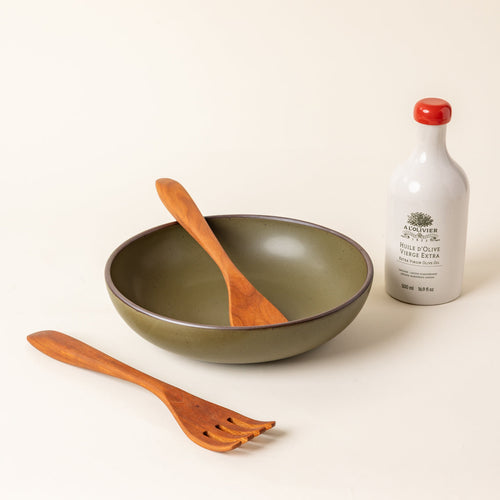 A large, shallow ceramic serving bowl in a matte olive green color featuring an unglazed rim, paired with a bottle of olive oil and two wooden serving utensils, against a cream background.