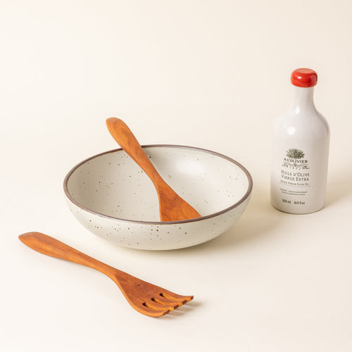 A large, shallow ceramic serving bowl in a matte warm off-white color featuring an unglazed rim, paired with a bottle of olive oil and two wooden serving utensils, against a cream background.