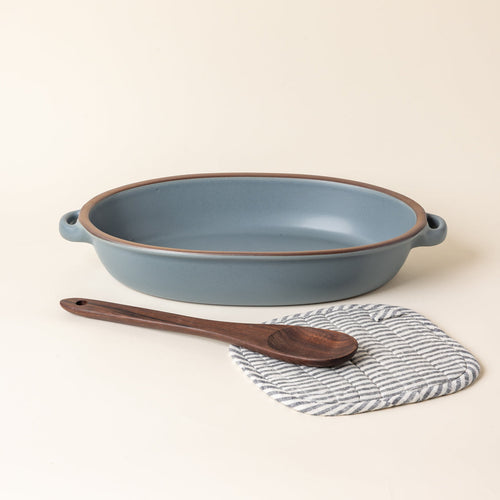 A large ceramic oval baker in a blue-grey color with a wood spoon and striped pot holder.