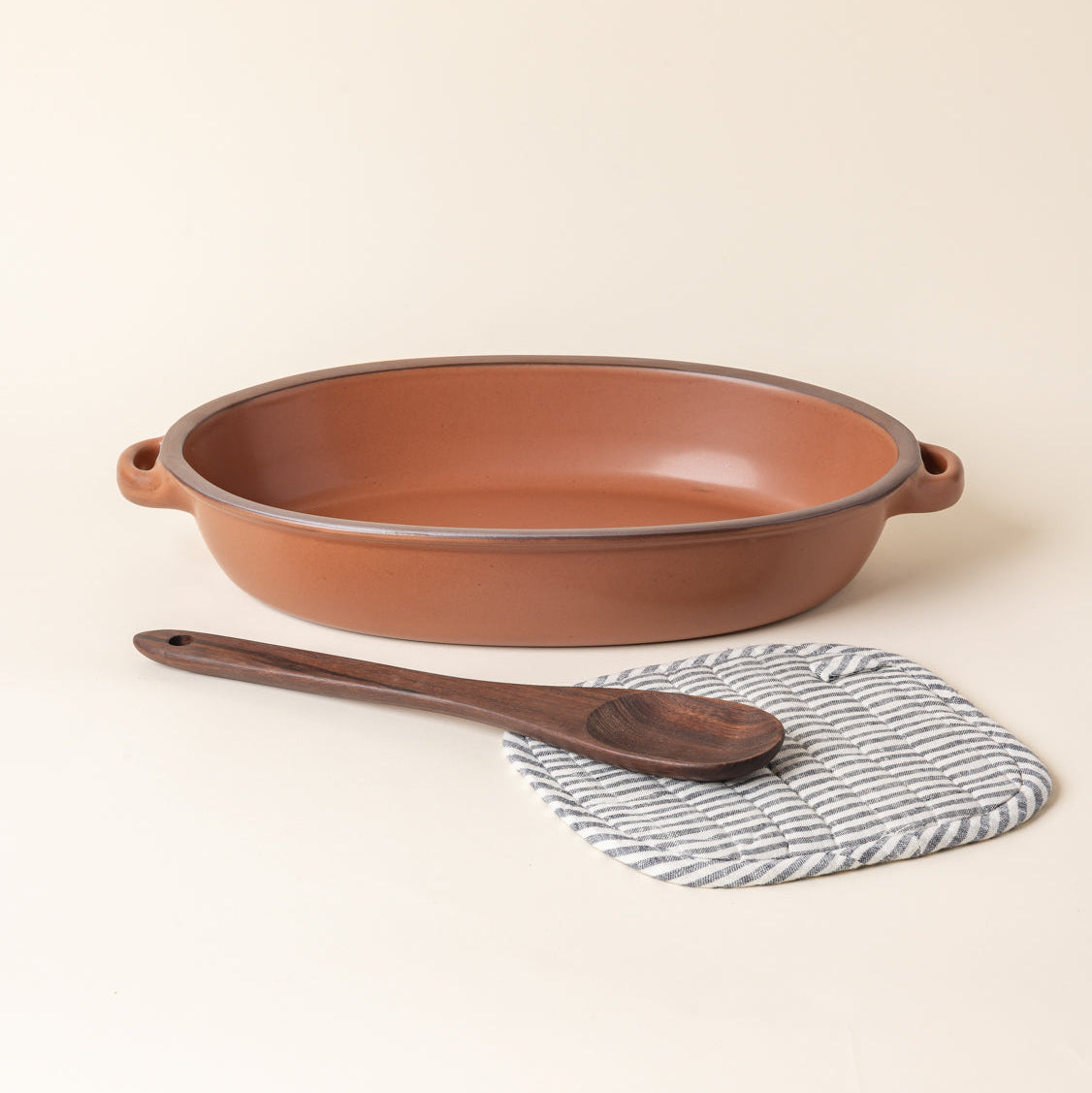 A large ceramic oval baker in a cool terracotta color with a wood spoon and striped pot holder.