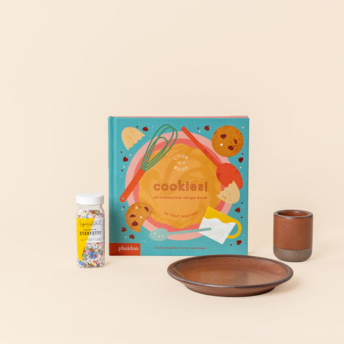 A colorful book titled 'cookies!', a bottle of rainbow sprinkles, and a matching tiny cup and cake plate in a cool terracotta color with unglazed rims.