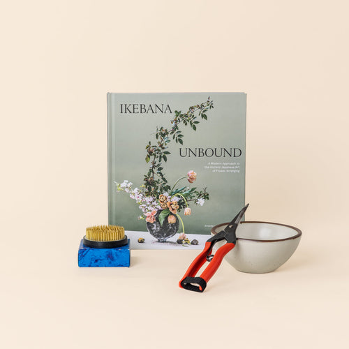 A book titled 'IKEBANA UNBOUND' standing upright, with a medium flower frog on a blue box, red-handled pruning shears, and a small warm off-white ceramic bowl arranged in front on a warm cream background.