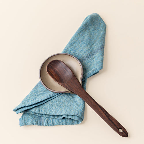 A ceramic spoon rest in a pale warm brown color with an unglazed rim, a blue kitchen towel, and a wooden kitchen spoon.