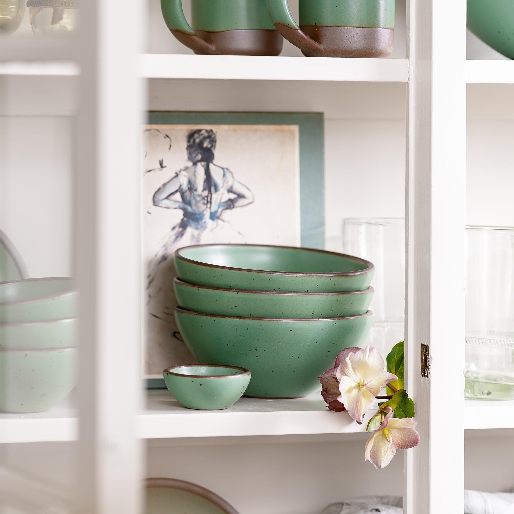 Stacked green ceramic bowls on a white shelf beside a framed sketch and a small flower, viewed through a cabinet frame.