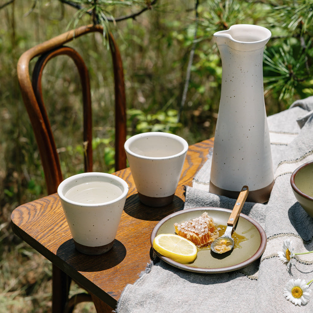 Ceramic carafe and cups in an off-white color on a wooden table outside.