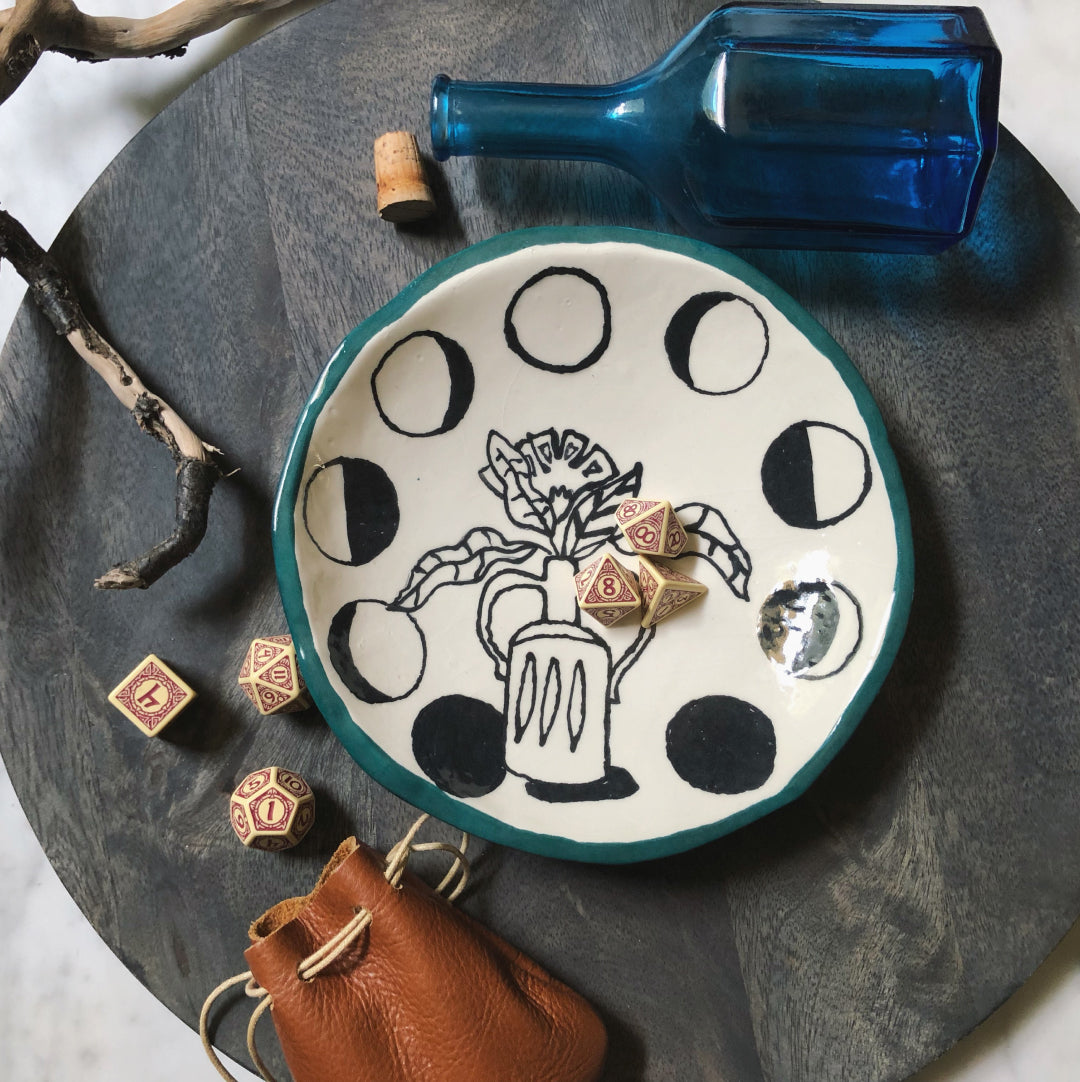 Decorative plate with moon phases, a bottle, and dice with a leather pouch on a dark surface.