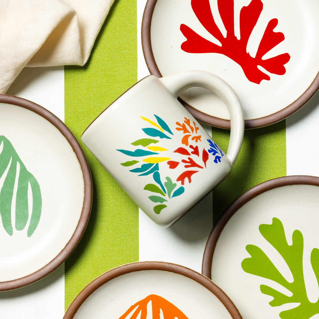 A full dip warm off-white mug with unglazed rim with colorful leaves decaled from Acanthes by Henri Matisse, styled with cake plates from The Matisse Collection.