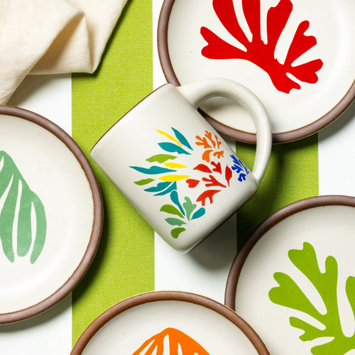 A full dip warm off-white mug with unglazed rim with colorful leaves decaled from Acanthes by Henri Matisse, styled with cake plates from The Matisse Collection.