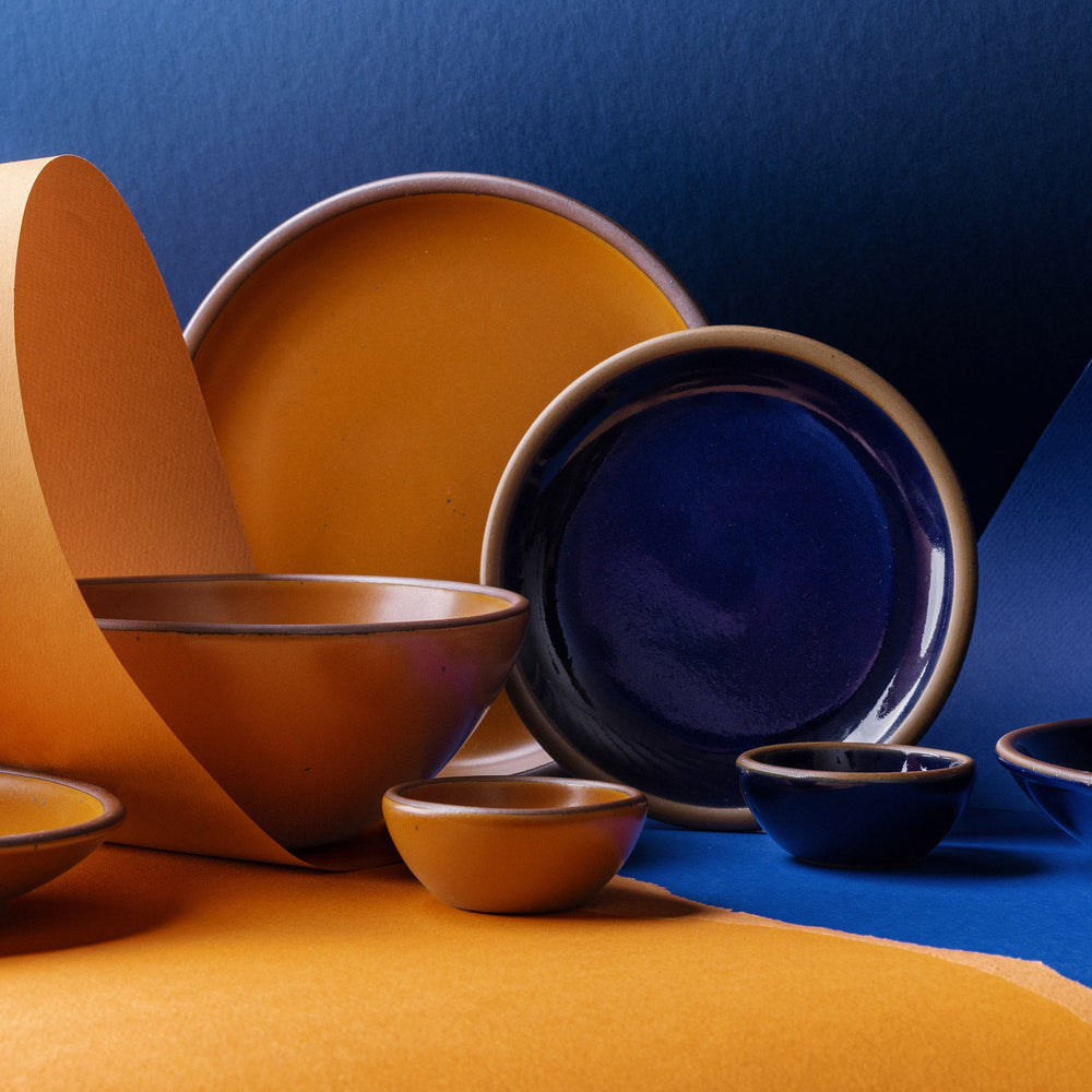 A styled studio arrangement of ceramic tableware in two contrasting glazes: a dark glossy blue and a warm earthy orange. The set includes plate and bowls, some nested together and others displayed upright, set against a split blue and orange paper background.