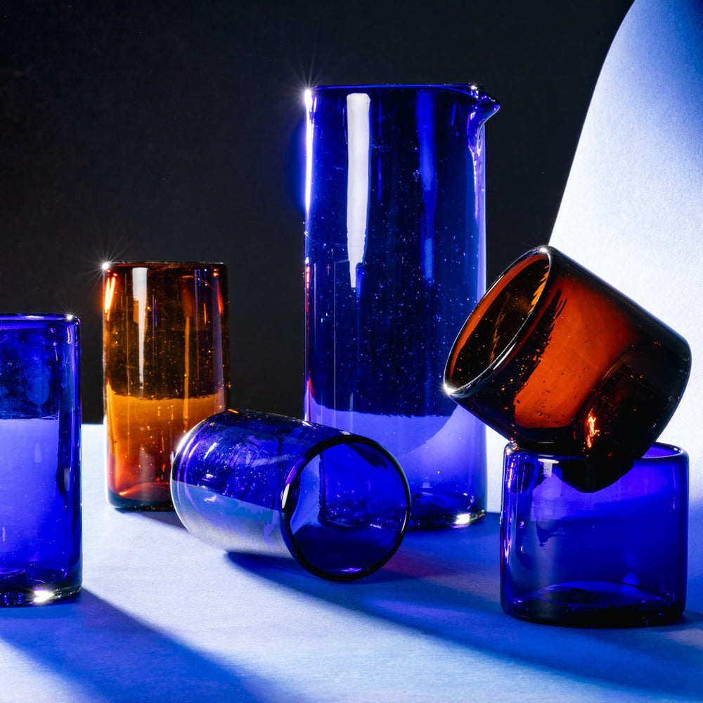 Short and tall tumblers and a pitcher in clear recycled glass in cobalt and dark amber colors.