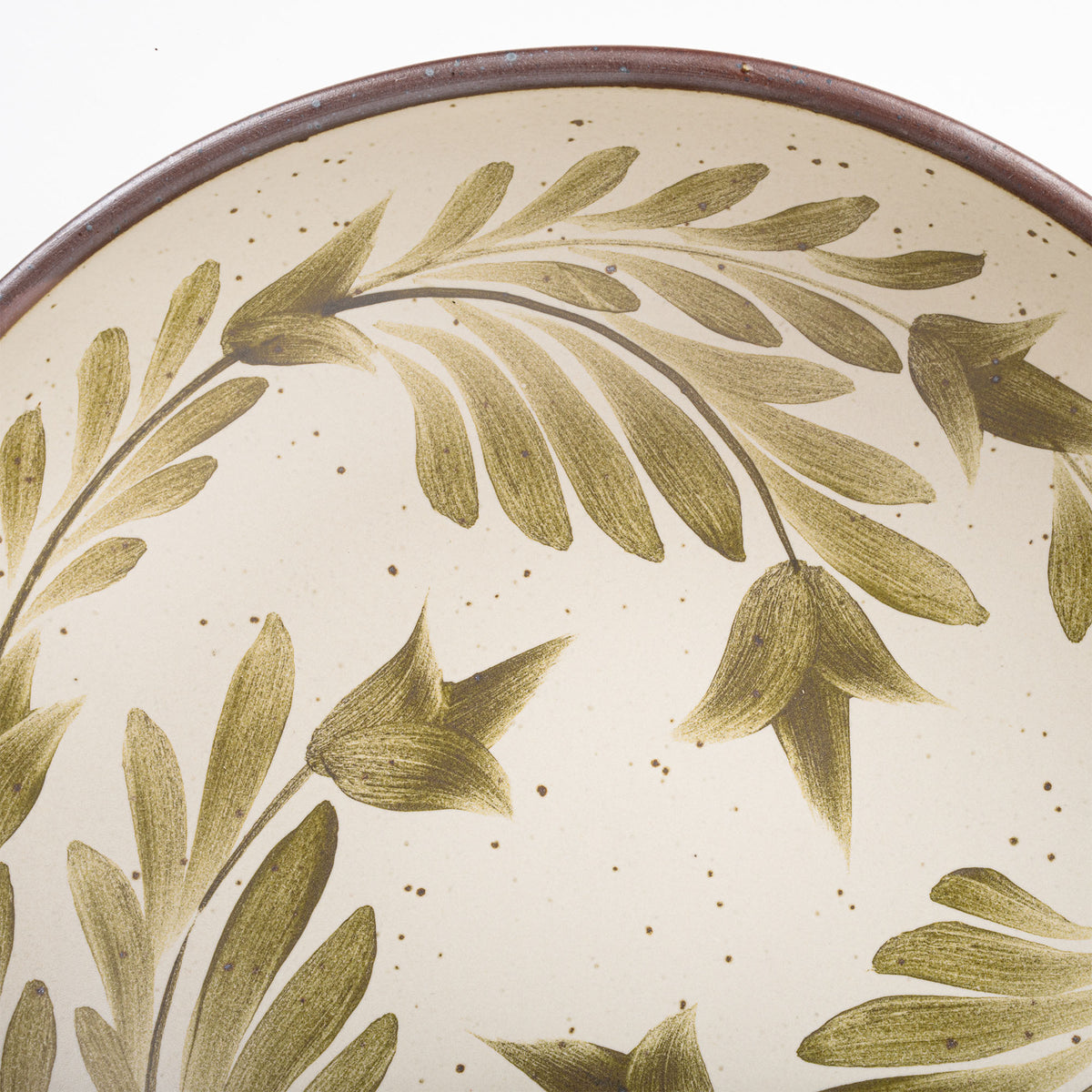 A closeup of a large shallow serving ceramic bowl in a warm, tan-toned, off-white color featuring iron speckles and an unglazed rim, with handpainted greenery in an olive green color inside the bowl.