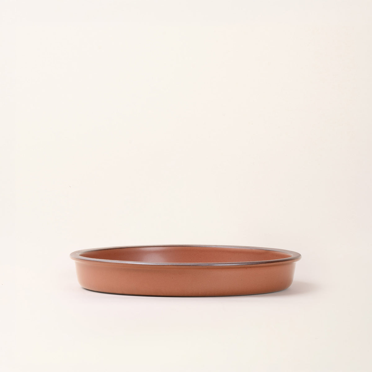 A straight on view of a shallow round ceramic baking dish in a matte cool terracotta color featuring an unglazed rim