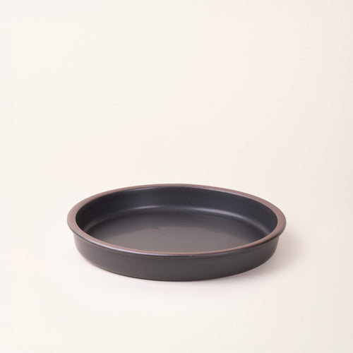 A shallow round ceramic baking dish in a matte graphite black color featuring an unglazed rim, photographed at an angle against a cream background.