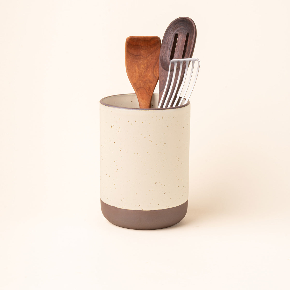 A cylindrical ceramic utensil crock and an unglazed base, in a warm off-white color, with various kitchen utensils inside