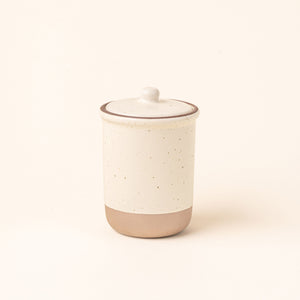 A tall ceramic lidded jar in a warm off-white color with an unglazed base.