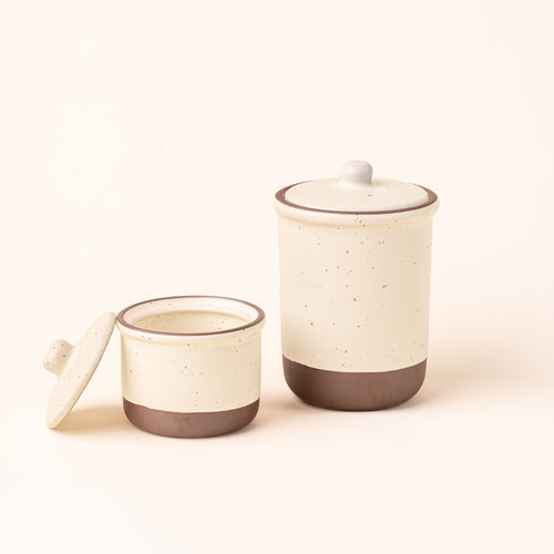 A small and tall ceramic lidded jars in a warm off-white color with an unglazed base.