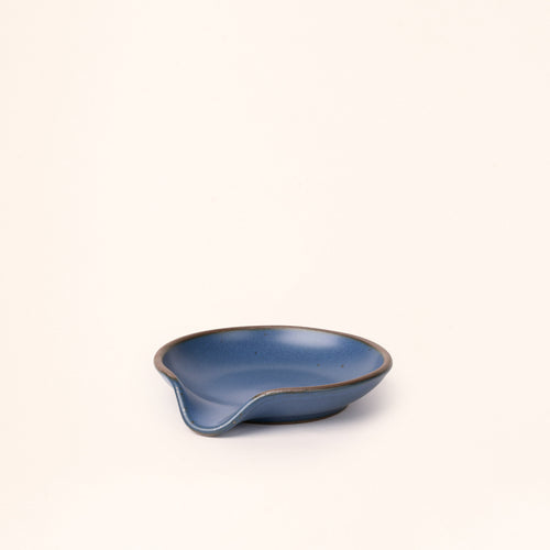 A side view of a ceramic spoon rest with a curved notch in a grounded blue color with an unglazed rim.