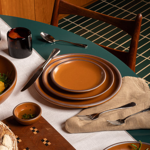 Earthy orange ceramic plates with amber glass, silver utensils and glassware arranged on a green dining table.
