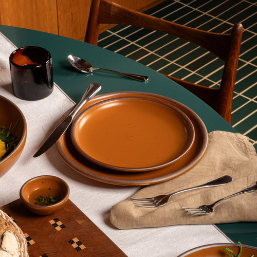 A styled dining table setting featuring a stack of earthy orange plates, silver flatware, a dark amber glass candle holder, and a beige linen napkin on a teal table runner.