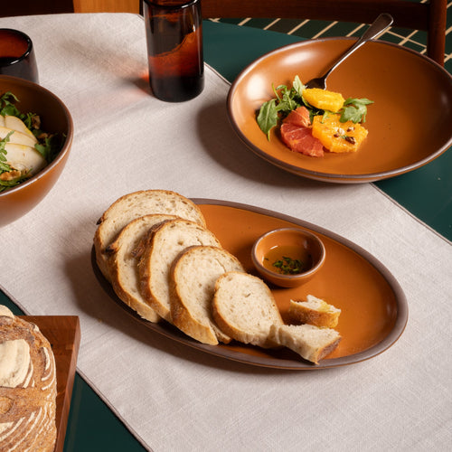 Medium oval ceramic platter in an earthy orange color with sliced bread and a small bowl of olive oil on a dark green table.