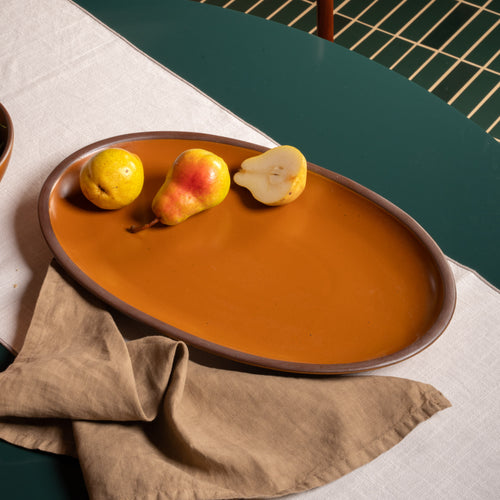 Large oval ceramic platter in an earthy orange with whole and halved pears on a dark green table.