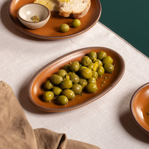 Small oval ceramic serving dish in an earthy orange color filled with green olives and lemon peel on a dark green table.