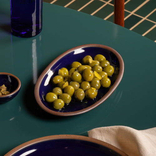 Small oval ceramic serving dish in a glossy dark blue color filled with green olives and lemon peel on a dark green table.
