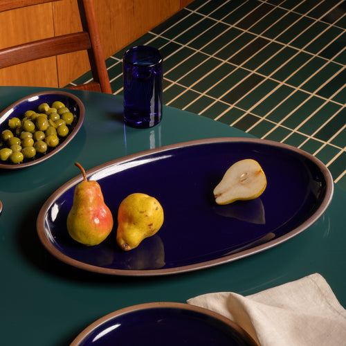 Large oval ceramic platter in a glossy dark blue with whole and halved pears on a dark green table.