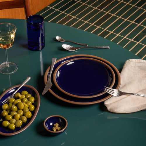 A styled dining table setting featuring a stack of ceramic plates in dark glossy blue with a brown rim, silver cutlery, a glass of white wine, a blue tumbler, a bowl of green olives, and a folded cream napkin on a teal table.