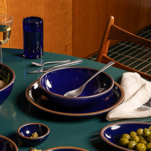 Glossy dark blue ceramic shallow bowl and dinner plate with silver utensils on a green dining table, surrounded by blue serving dishes.
