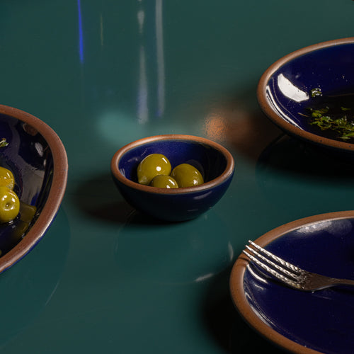 Tiny glossy dark blue ceramic bowl filled with green olives on a dark green table.